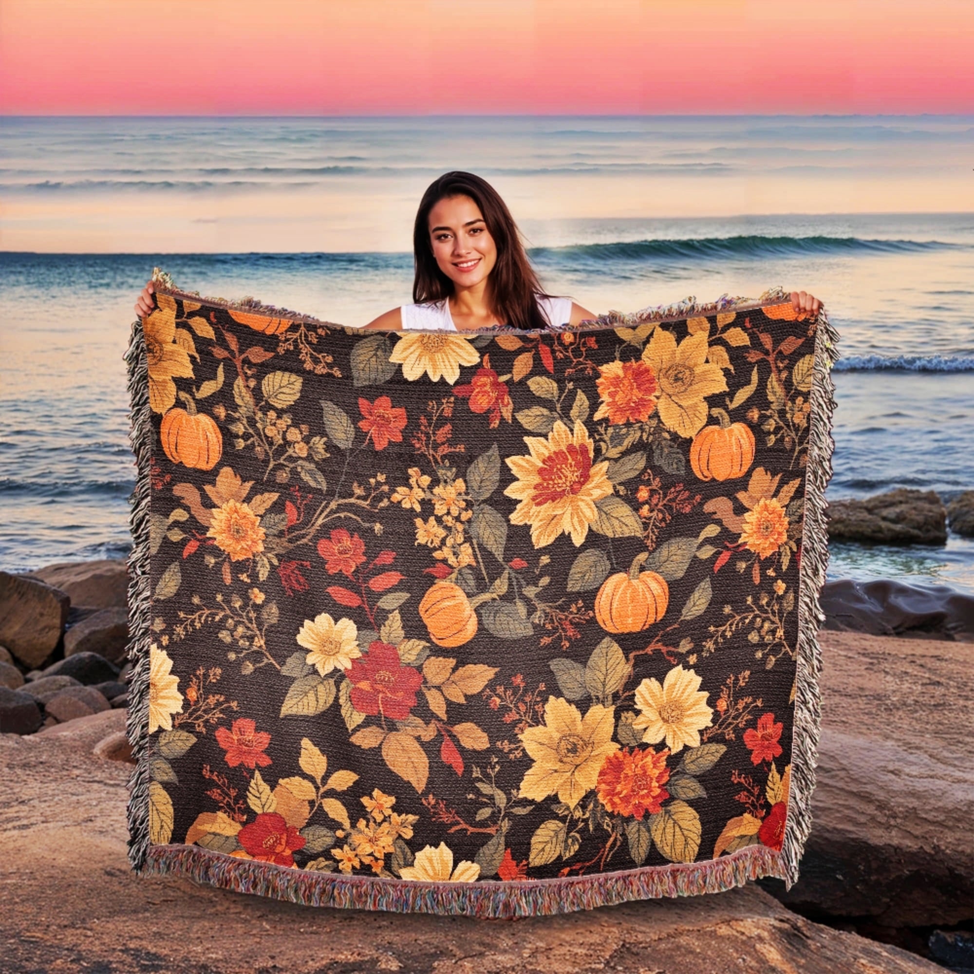Woman holding a floral and pumpkin-patterned blanket by the ocean at sunset