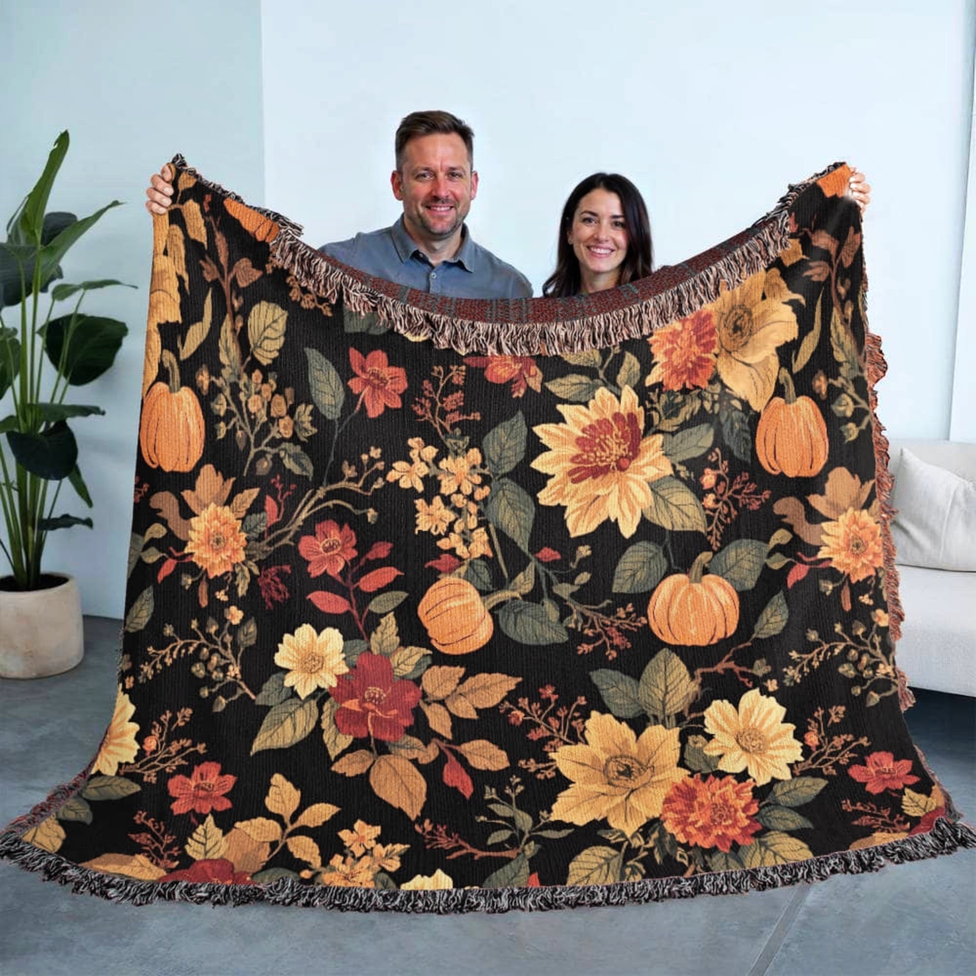 Two people holding a floral and pumpkin patterned blanket indoors.